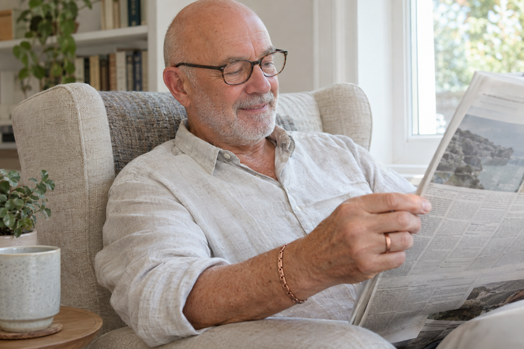 Richard reading a newspaper by the window, wearing a copper ring and bracelet